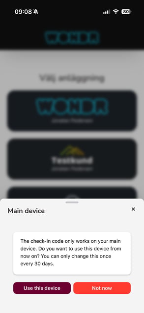 Login prompt — switch main device Wondr app modal titled "Main device," appearing over a blurred facility-picker screen. It reads: "The check-in code only works on your main device. Do you want to use this device from now on? You can only change this once every 30 days," with "Use this device" and "Not now" buttons.